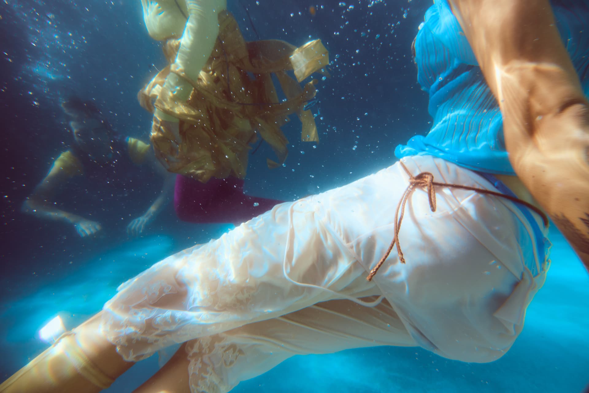 GOODBYE SUMMER — close-up of white lace trousers drifting underwater with figures behind
