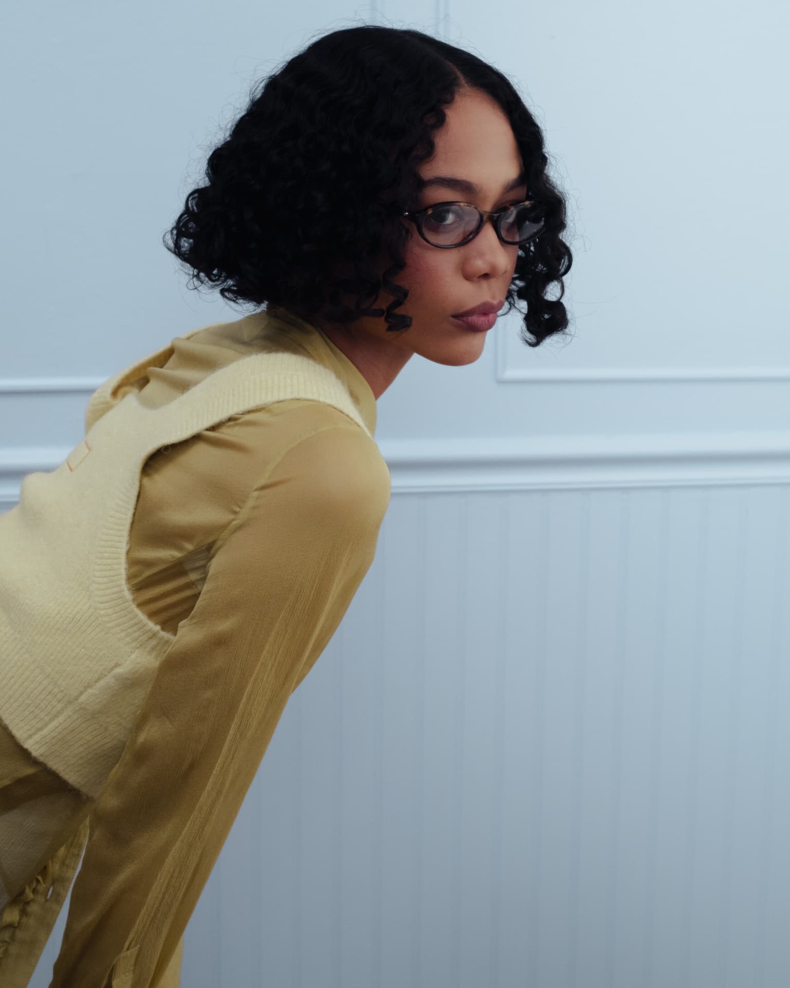 Bonnie Clyde — woman with short curly hair and dark-rimmed glasses in a yellow sheer top and knit vest