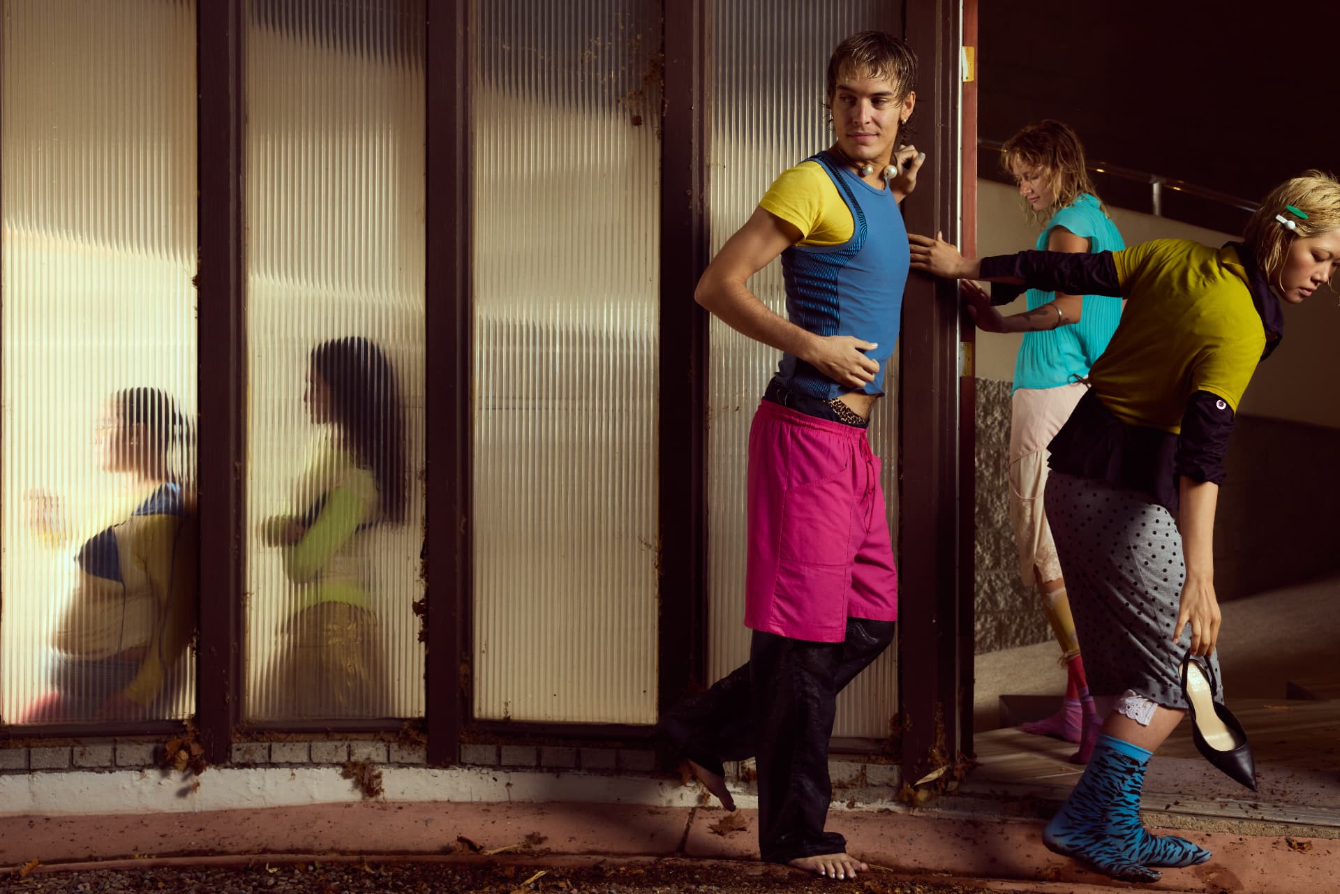 GOODBYE SUMMER — three people in bright wet outfits by an industrial doorway with silhouettes behind glass